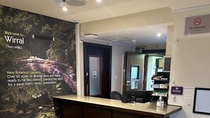 A reception area with a wooden reception desk, a 'No Smoking' sign, a 'Buzzer' sign, and a poster advertising 'Ness Botanical Gardens'. - Premier Roomz Two Mills (Ellesmere Port)