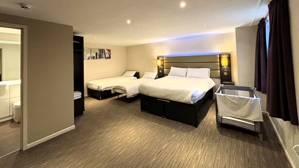 A hotel room with two beds, a small seating area, a bathroom, and a view of the cityscape. - Premier Roomz Two Mills (Ellesmere Port)