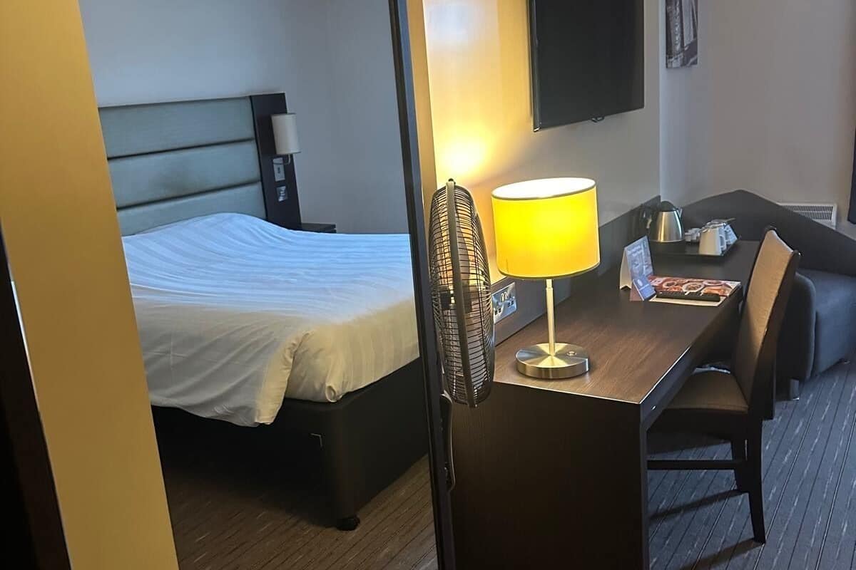 Quarto de hotel com uma cama, uma escrivaninha com luminária, uma cadeira e uma televisão de tela plana na parede.