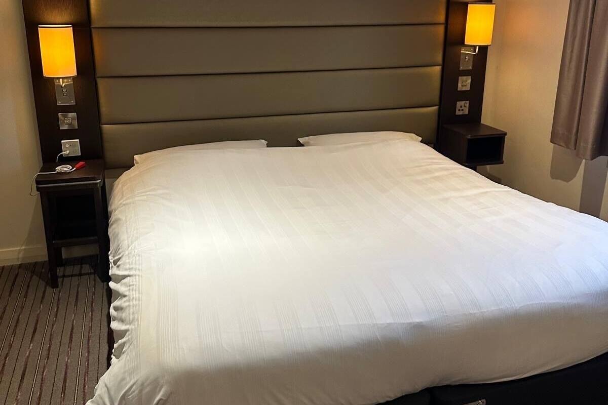 Quarto de hotel com uma cama grande, duas mesas de cabeceira com luminárias, cabeceira com iluminação embutida e um tapete listrado.