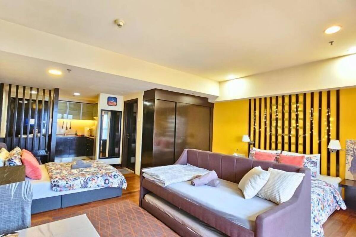 A modern hotel room with a sofa, a bed, a wardrobe, and a kitchen area visible through a glass partition.