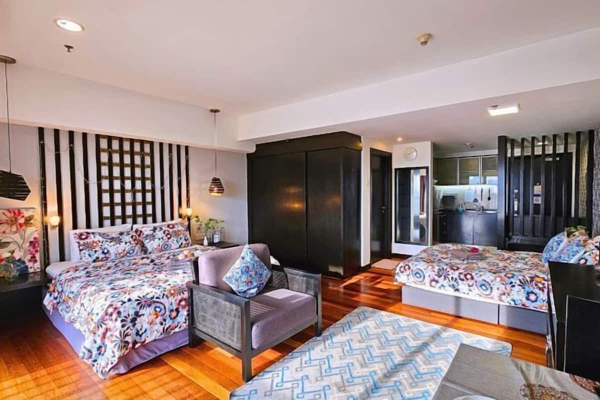 A modern hotel room with two beds, a sofa, a dining table, and a kitchen area.
