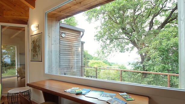 Un escritorio de madera con libros y una silla en una habitación con un ventanal grande que da a un jardín.
