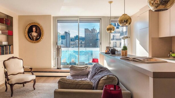 A modern living room with a sofa, a chair, a bookshelf, a large window with a city view, and a kitchen area with a counter and pendant lights. - AN ICONIC SPACE  IN AN ICONIC LOCATION  - AVENUE RAPP – NEXT TO EIFFEL   (Paris)
