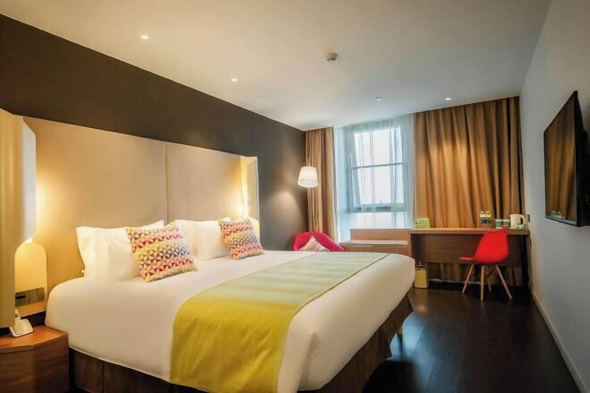 A hotel room with a large bed, a desk, a chair, and a TV on the wall.