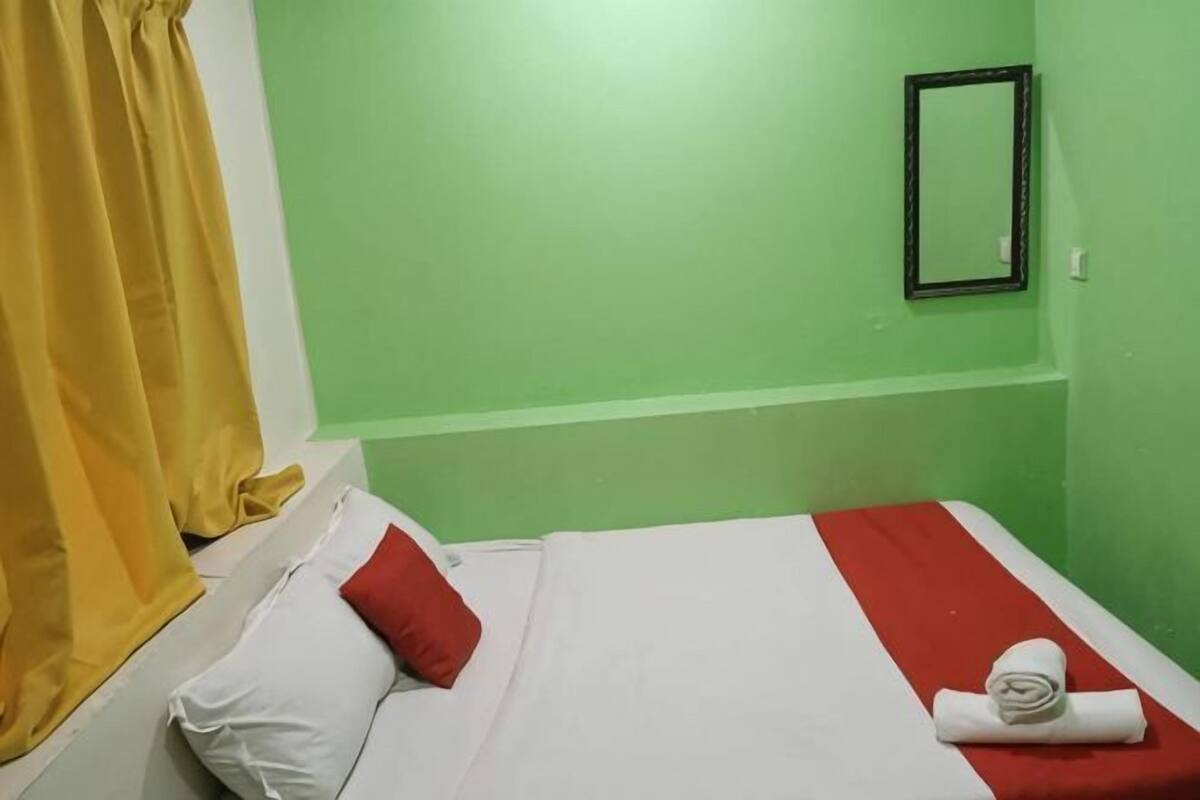 A room with a bed, a red and white bedspread, a white pillow, a red pillow, a folded towel, a yellow curtain, and a black-framed mirror on the wall.