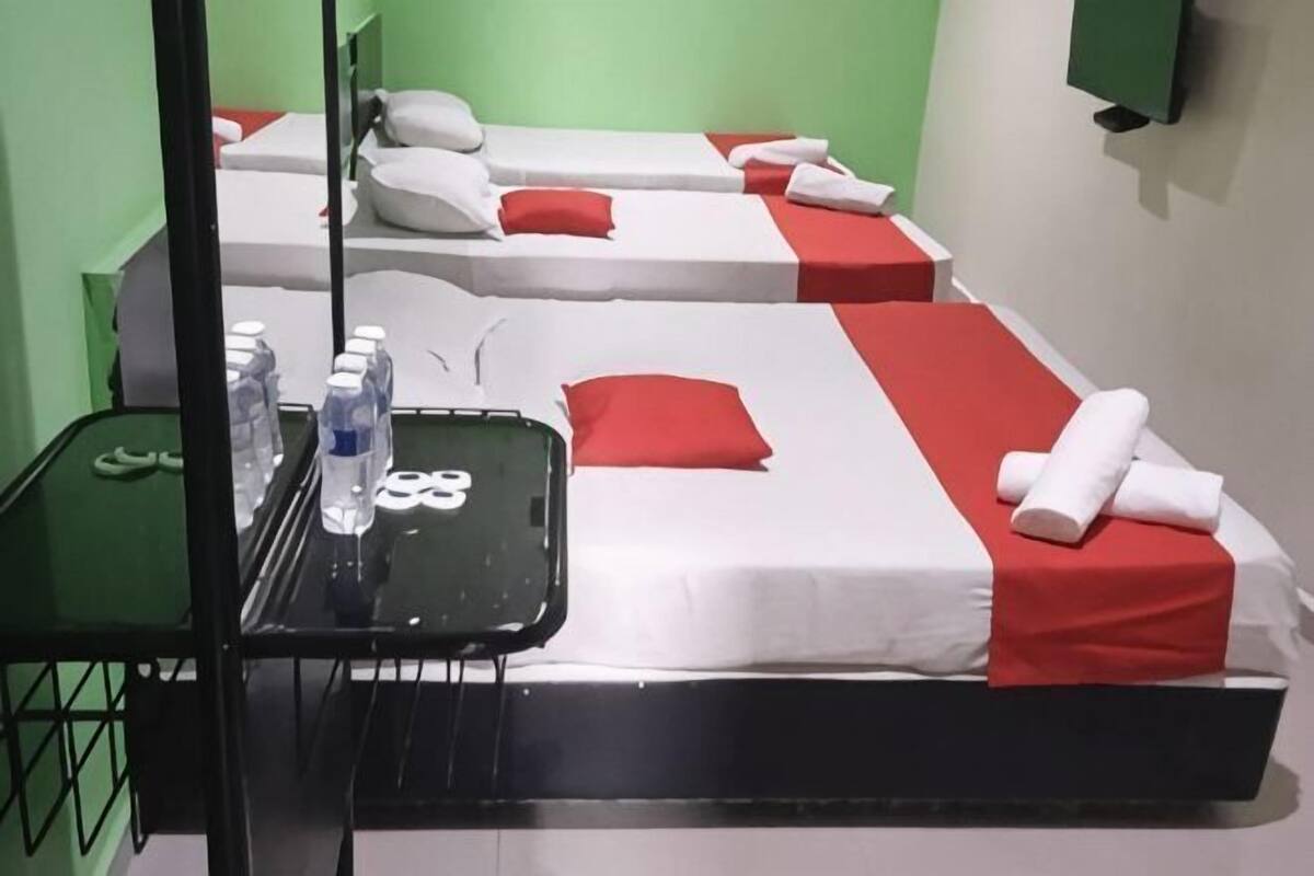 A hotel room with three beds, each with a red pillow and white bedding. There is a small table with a tray containing a bottle of water, a glass, and a pair of glasses.