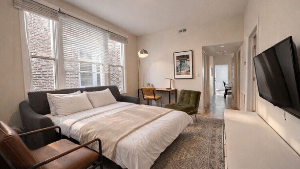 A bedroom with a bed, a desk, a chair, a TV, and a window with blinds. - Flexhome Wicker Park • Stylish 2BR • Sleeps 6 (Chicago)