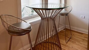 A dining area with a glass table and metal wire chairs, a window with blinds, and wooden flooring. - Flexhome Wicker Park • Stylish 2BR • Sleeps 6 (Chicago)