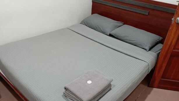 A neatly made bed with a grey bedspread and pillows, a folded grey towel, and a wooden headboard. - Evonne Budget Hotel (Tanah Rata)