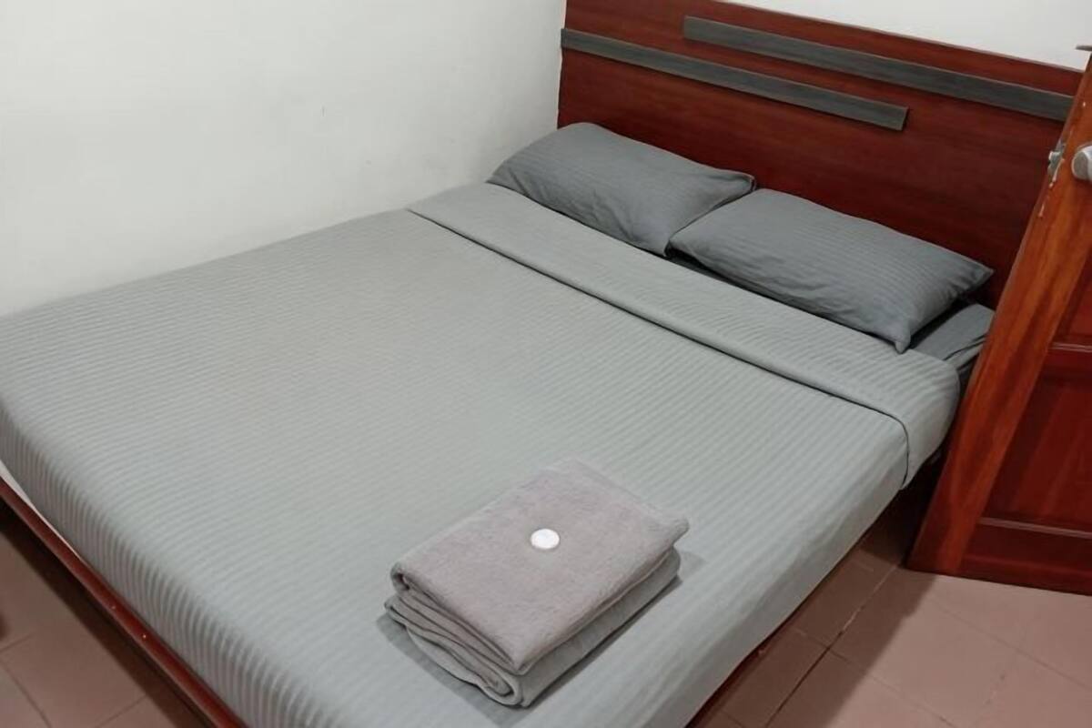 Un lit bien fait, avec une couverture et des oreillers gris, une serviette grise pliée et une tête de lit en bois.