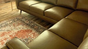 A modern living room with a brown leather sofa, a patterned rug, and a large window with blinds.