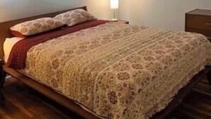 A bedroom with a wooden bed, a bedside table, a lamp, and a window with blinds.