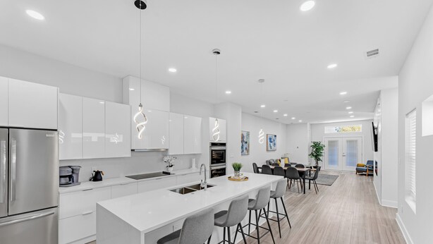 Una cocina moderna con armarios blancos, una isla central con taburetes de bar, electrodomésticos de acero inoxidable e iluminación empotrada en el techo.