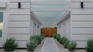 Un bâtiment moderne doté d’un passage central ouvert, bordé de deux murs blancs, qui mène à un portail en bois.