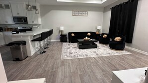 Un salon moderne avec un canapé noir, une table basse et un fauteuil noir. On y trouve également un coin cuisine avec des meubles blancs et un four à micro-ondes. Un ventilateur de plafond est visible, et un rideau noir est accroché à la fenêtre.