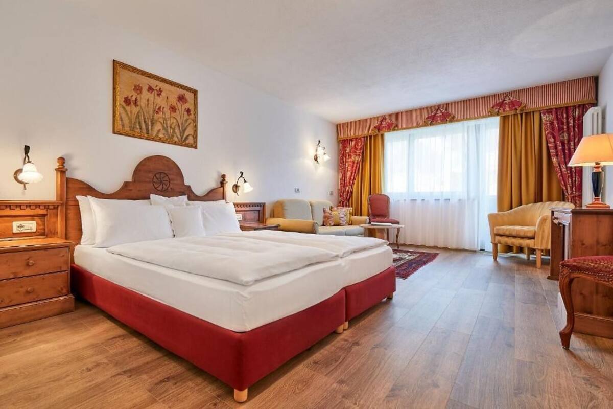 A hotel room with a large bed, a wooden nightstand, a desk, a chair, and a window with curtains.