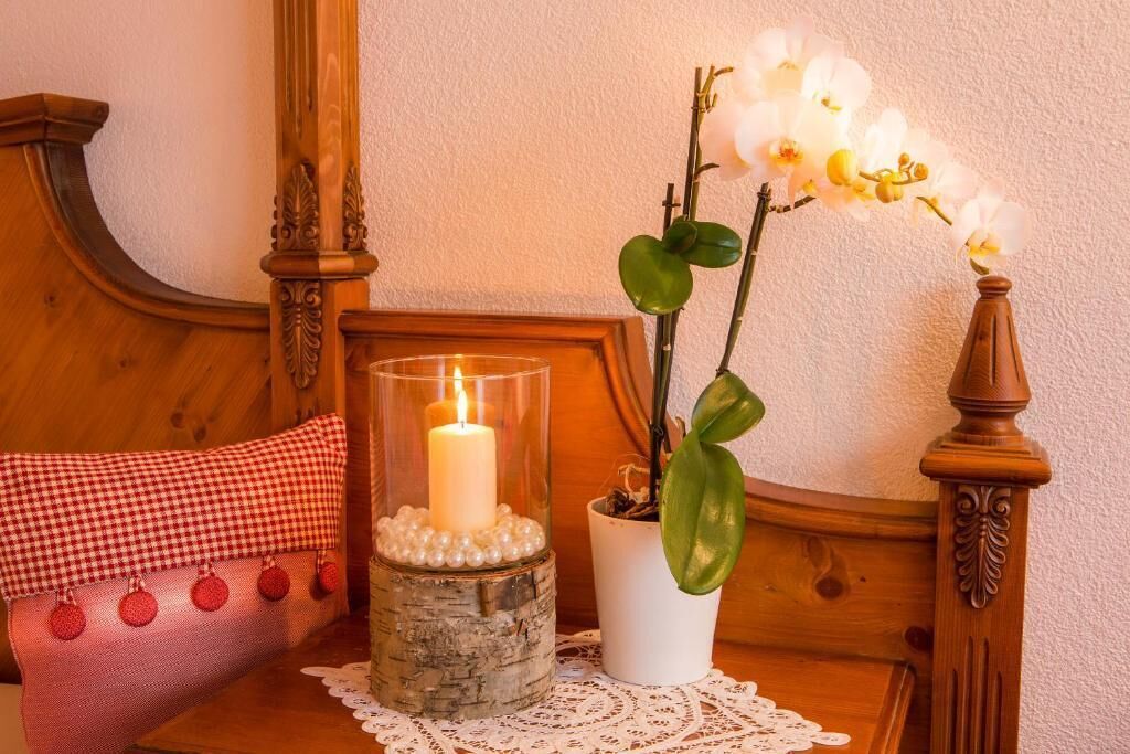 A wooden bed with a glass candle holder containing a lit candle, a checkered pillow, and a potted orchid plant.