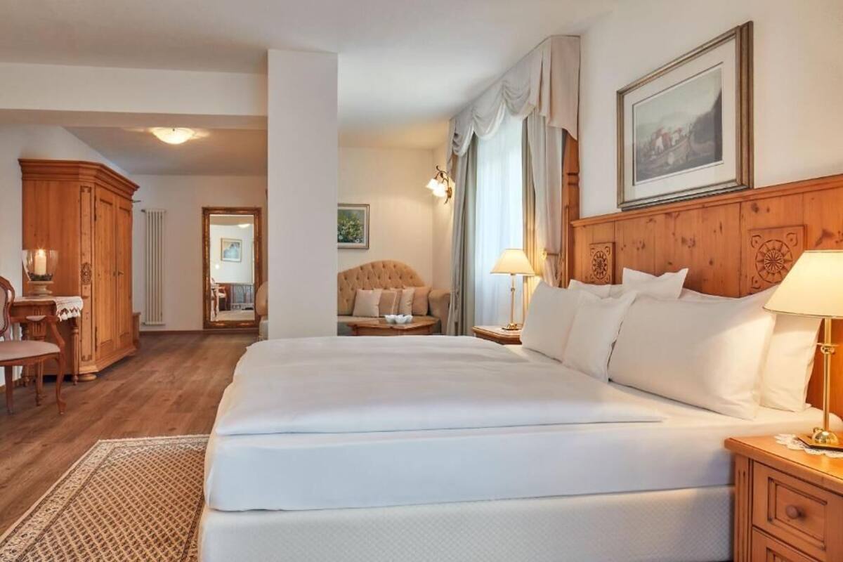 A hotel room with a large bed, wooden headboard, a wooden wardrobe, a small table with a lamp, and a view of the hallway.