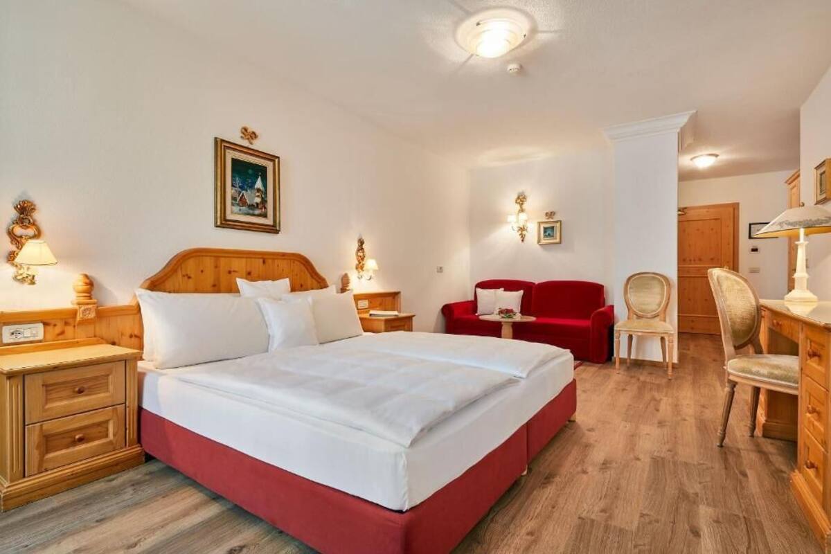 A hotel room with a large bed, wooden headboard, two bedside tables with lamps, a red sofa, a wooden chair, and a wooden dresser.