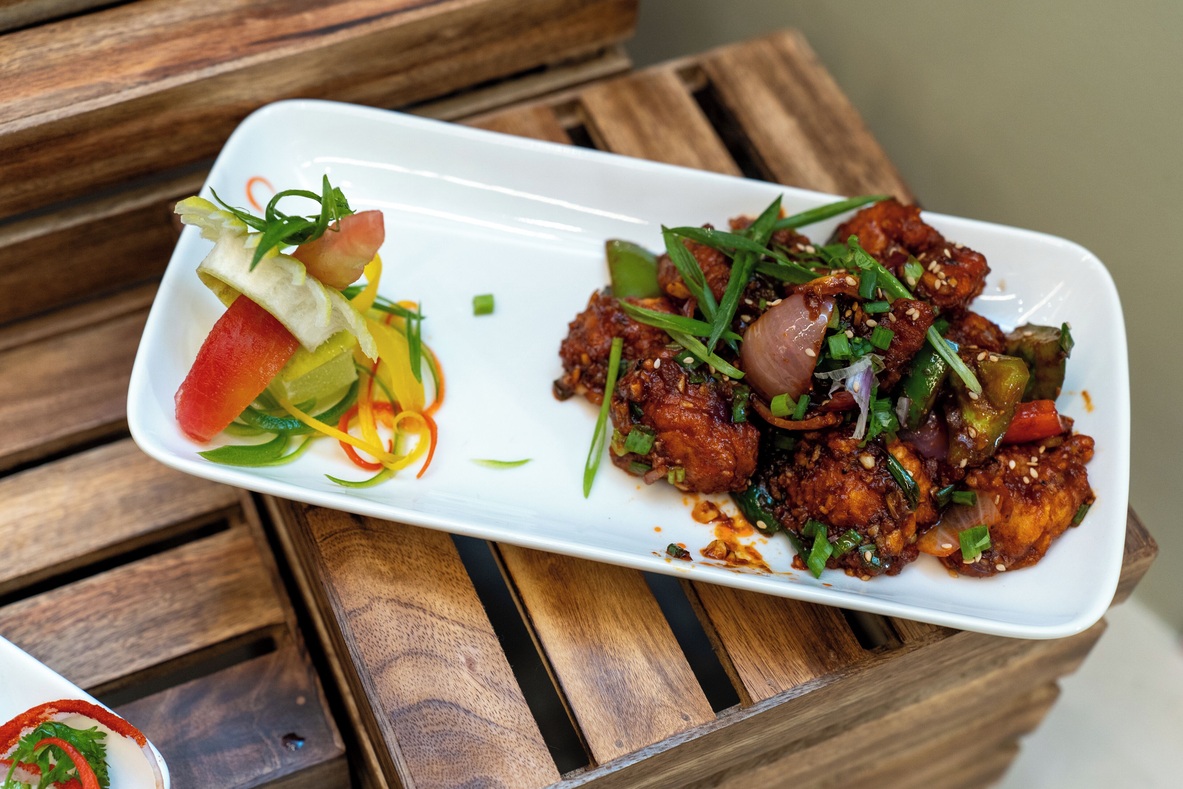 A rectangular white plate with a dish of stir-fried chicken and vegetables, garnished with green onions and sesame seeds, on a wooden table.