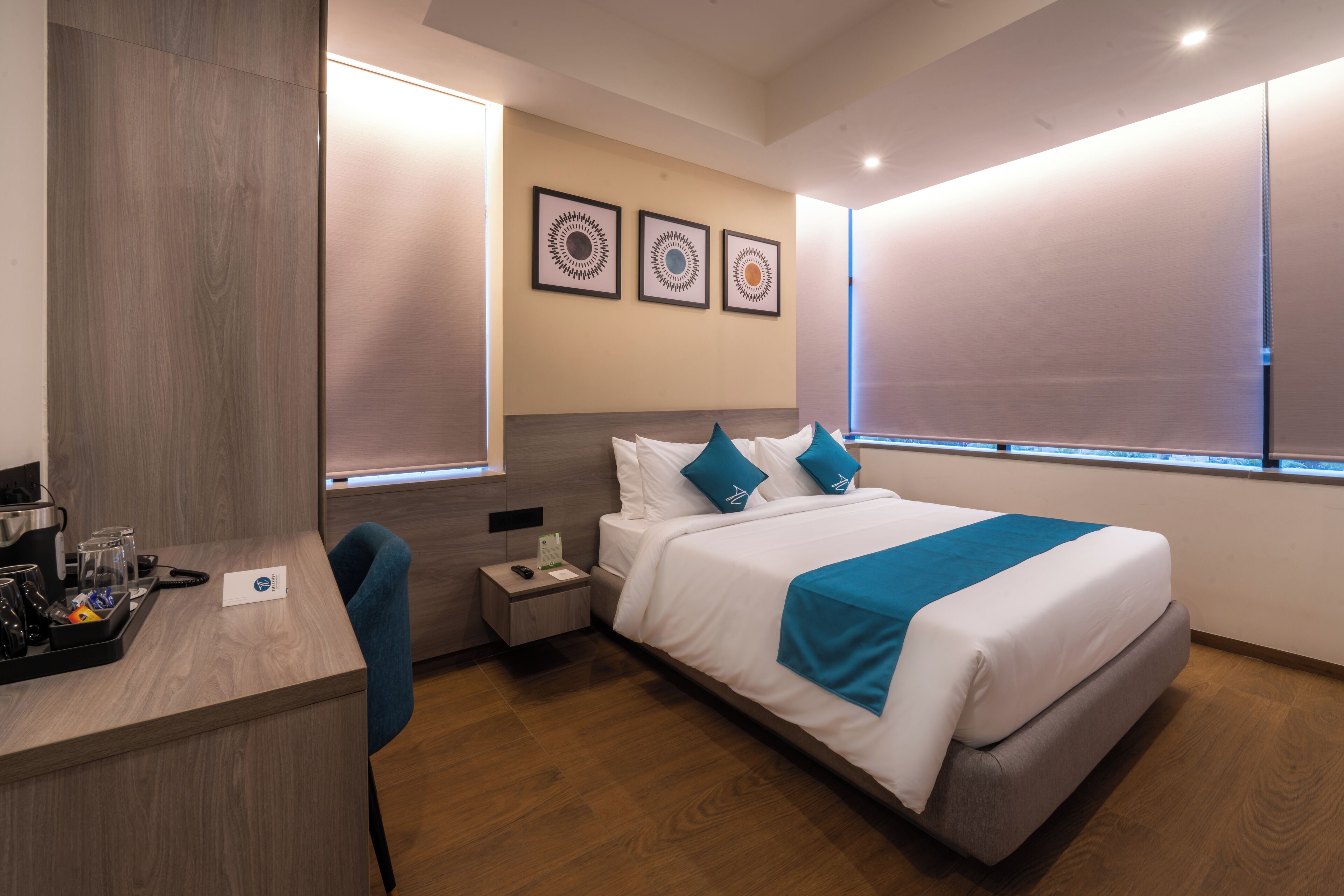 A modern hotel room with a bed, bedside tables, a desk with a chair, and artwork on the wall.