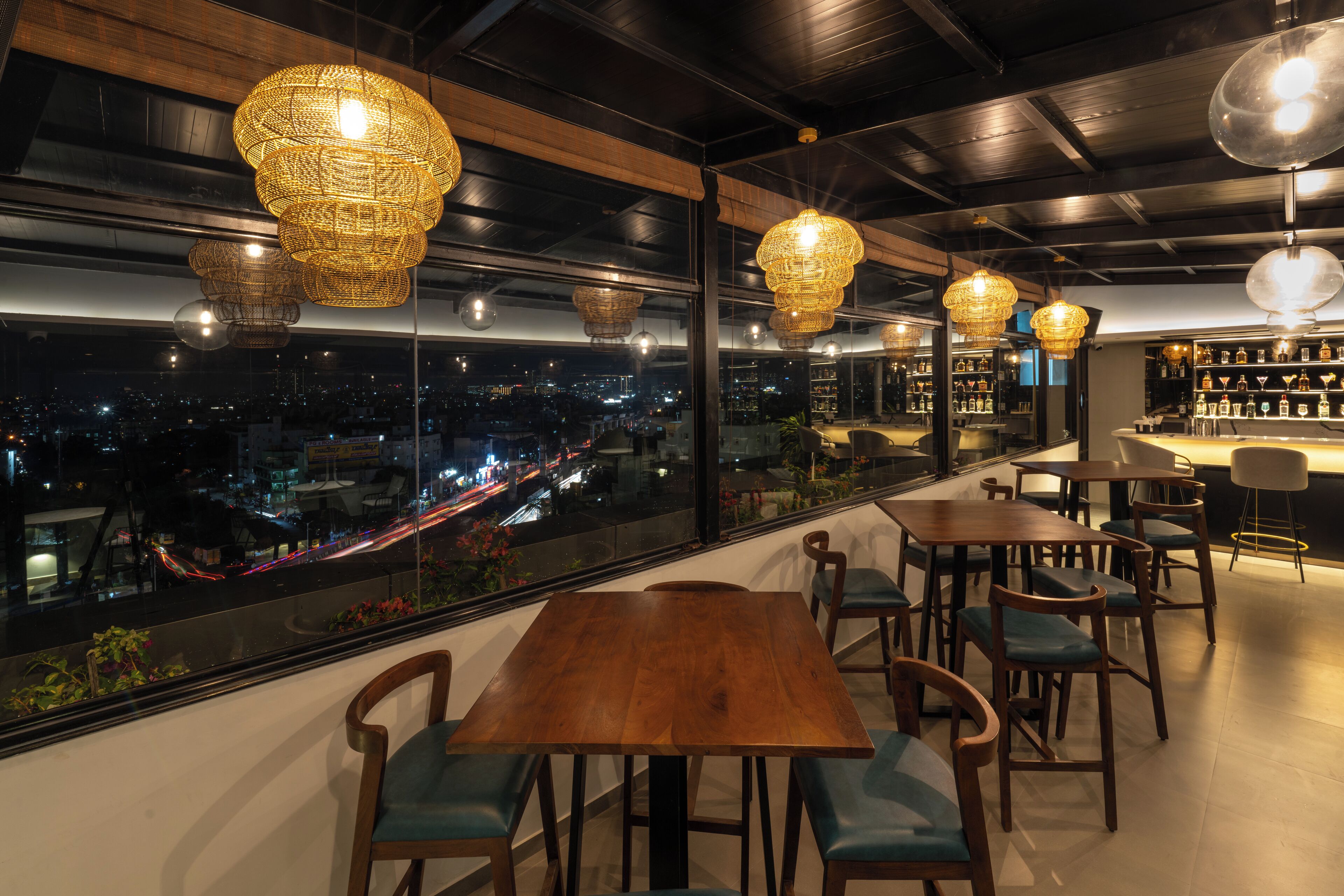 Un restaurante con vistas a la ciudad de noche, con mesas y sillas de madera, y lámparas colgantes de mimbre.