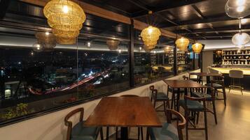 Un restaurante con vistas a la ciudad de noche, con mesas y sillas de madera, y lámparas colgantes de mimbre.