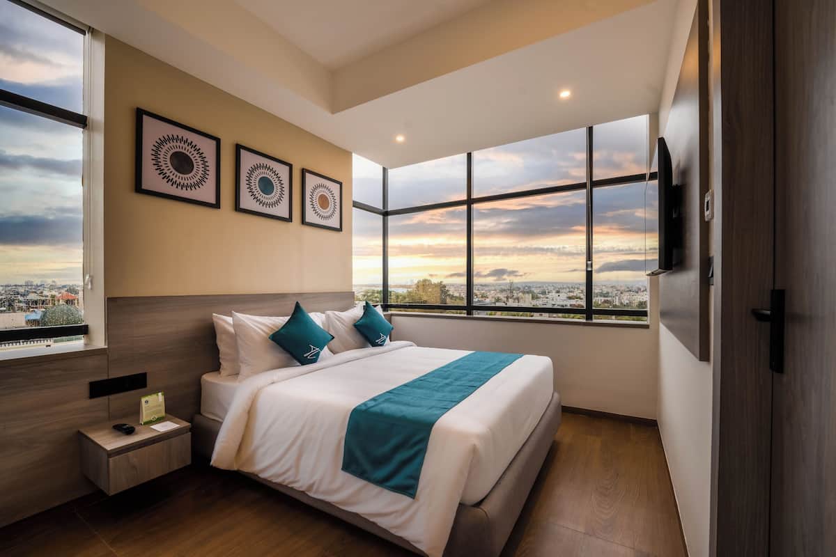 A modern hotel room with a large bed, a nightstand, a TV mounted on the wall, and a window with a city view.