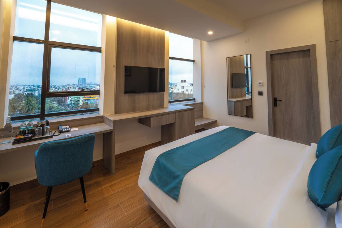A modern hotel room with a large bed, a desk with a TV, a chair, and a view of the city.