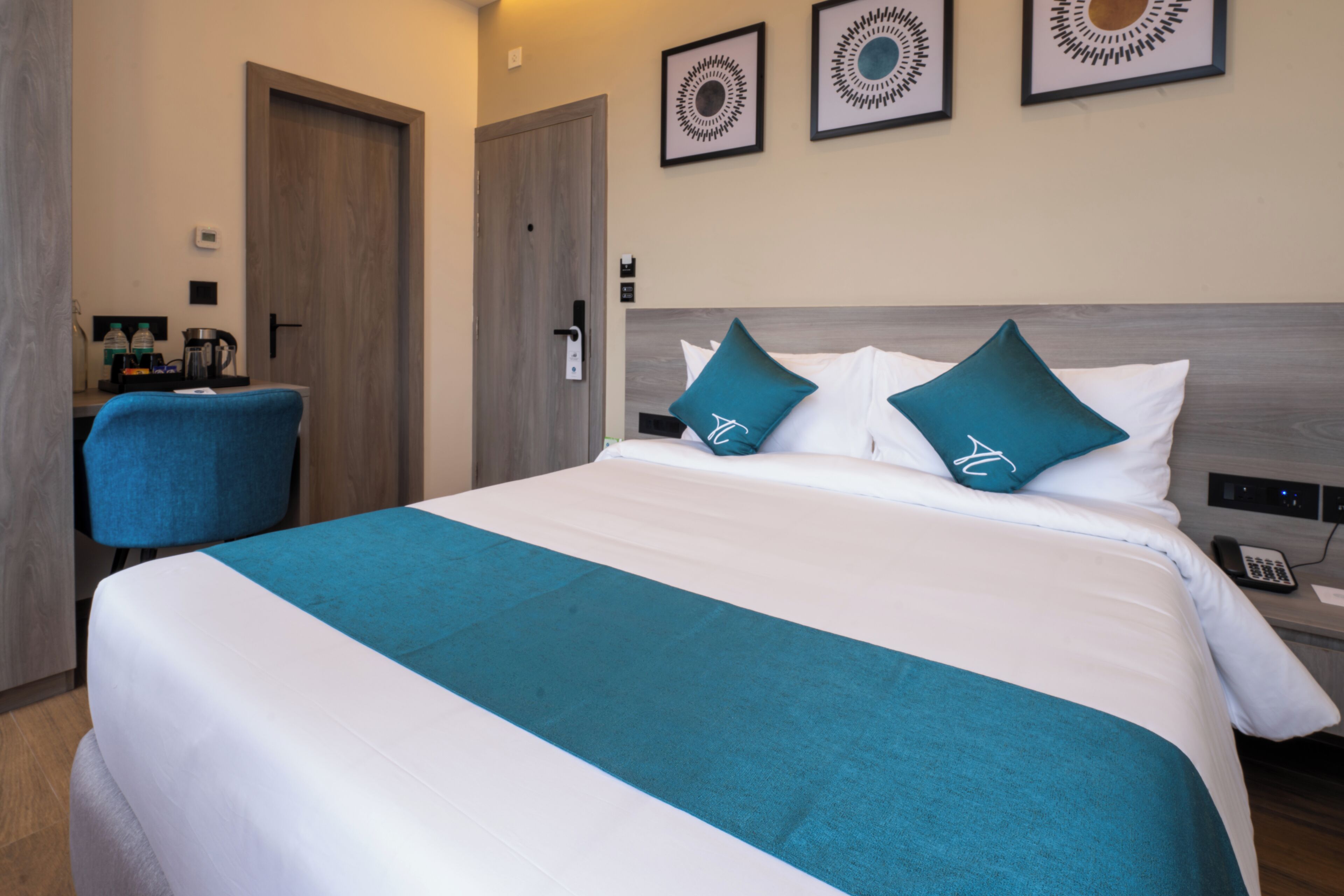 A hotel room with a large bed, two teal pillows, a wooden headboard, a desk with a chair, and a coffee maker.