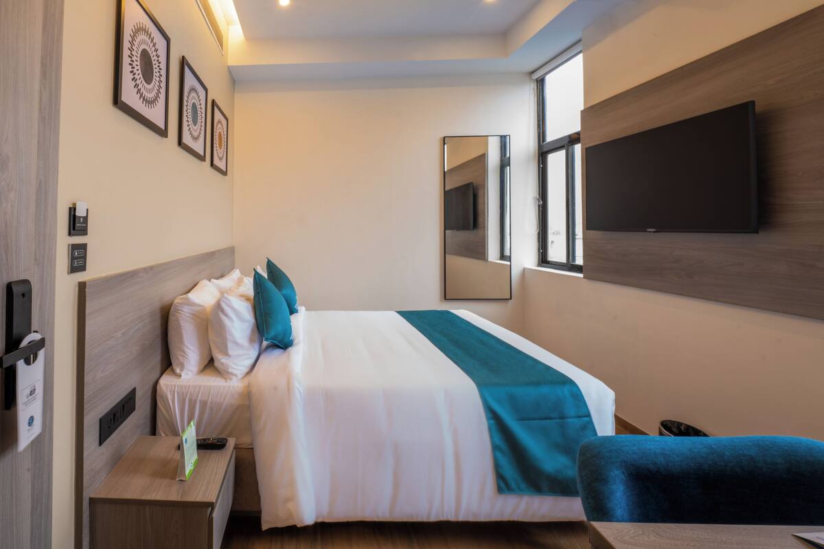 A hotel room with a bed, a desk, a chair, a TV mounted on the wall, and framed artwork on the wall.