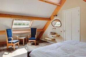Habitación con techo inclinado, vigas de madera, tragaluces, una cama, dos sillas, una mesita, un espejo y una estantería con un cuenco.