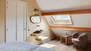 Un dormitorio con tragaluz, techo de vigas de madera, cama, silla y una mesita con un cuenco encima.