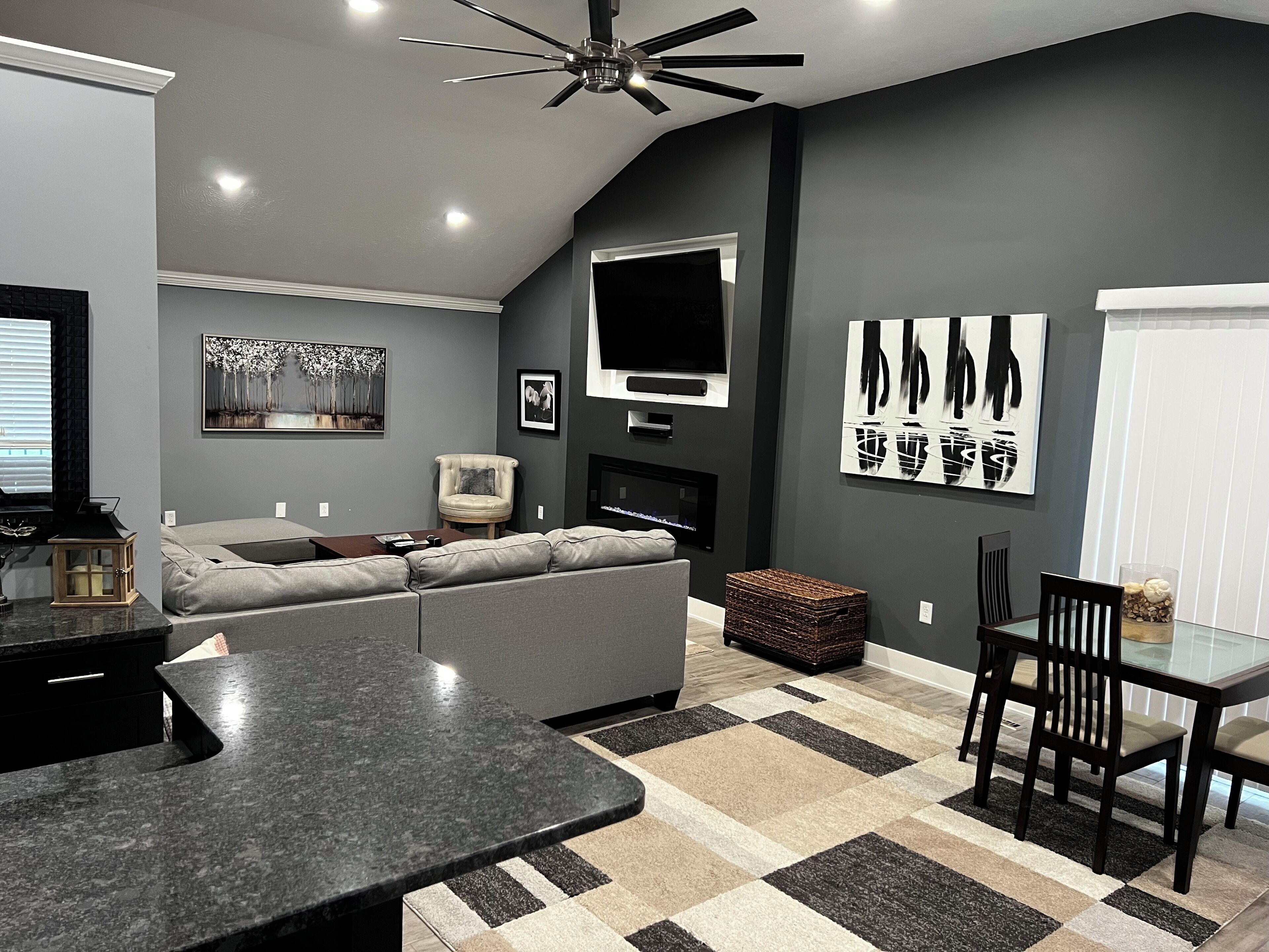 A modern living room with a sectional sofa, a flat-screen TV, artwork on the walls, a fireplace, and a dining area with a glass table and chairs.
