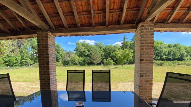 Un patio al aire libre con una mesa y sillas de vidrio, rodeado de muros de ladrillo y un techo de madera, con vistas a un campo de césped y árboles.