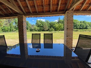 An outdoor patio with a glass table and chairs, surrounded by brick walls and a wooden ceiling, overlooking a grassy field and trees.