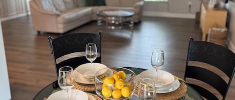 A dining area with a glass table set for four, surrounded by chairs, and a kitchen in the background.