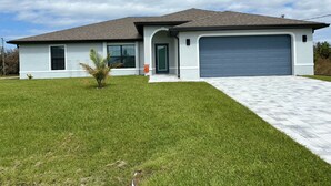 A modern two-story house with a grey garage door, a small front yard with grass and a palm tree, and a cloudy sky.