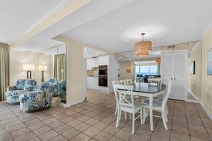 A spacious living room with a large mirror, a dining area with white chairs, and a kitchen with white cabinets. - Shoreline Towers 1081 (Destin)