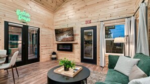 A cozy wooden interior with a green sofa, a round coffee table with a plant, a TV mounted on the wall, and an exit sign. - Lux Sauna, Dome, Stargazing Net, Putting Green (Sevierville)