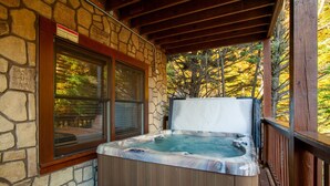 Un mur extérieur en pierre, une terrasse en bois, un jacuzzi et une fenêtre donnant sur une zone boisée.