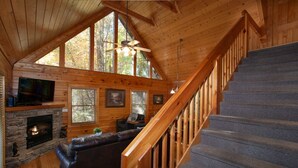 L’intérieur d’une cabane avec une cheminée en pierre, des murs en bois, un escalier et une grande fenêtre.