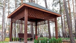 Un gazebo in legno con tetto a graticcio, panche e base in pietra, circondato da alberi e piante.