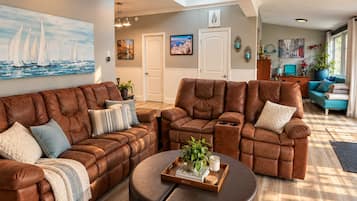 A spacious living room with a brown leather sectional sofa, a round ottoman, and a dining table with a tray of books and a candle.
