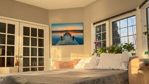 A bedroom with a bed, a large window, a painting of a sunset, and a door with glass panels. - Sauna, Pizza Oven, BBQ, Easy Lake Access & Parking for Trucks & Trailers (Winnipeg Beach)