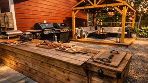 An outdoor barbecue area with a wooden grill, a wooden table with food and drinks, and a wooden bench.