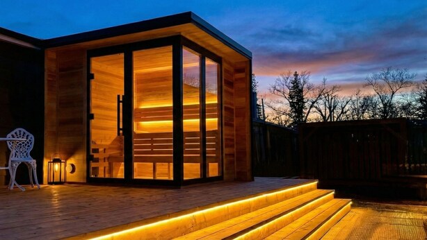 Un bâtiment moderne en bois, doté d’une entrée avec une porte en verre, d’un fauteuil en osier, d’une petite table et d’une terrasse en bois avec des marches éclairées.