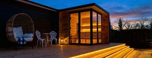 Un edificio moderno de madera con puerta de vidrio, una silla de mimbre, una mesita y una terraza de madera con escalones iluminados.