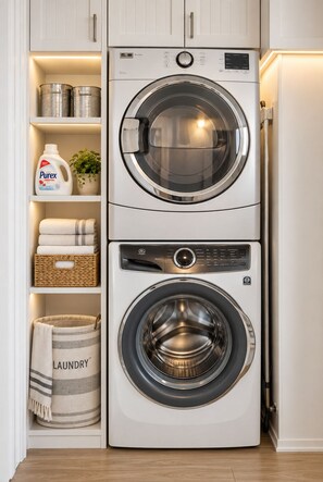 A compact, modern laundry room with two stacked front-loading washing machines, a shelf with laundry supplies, and a basket.